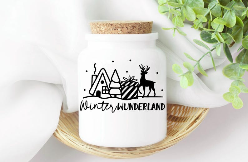 Puede incluir: Tarro de cer&aacute;mica blanco con tapa de corcho y un dise&ntilde;o de invierno de cuento de hadas en blanco y negro. El dise&ntilde;o incluye una casa, un &aacute;rbol, un regalo y un reno. El texto "Winter Wonderland" est&aacute; impreso debajo del dise&ntilde;o.