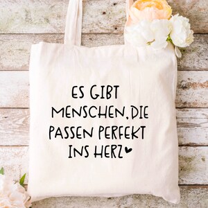 Puede incluir: Una bolsa de tela blanca con texto negro que dice "Es gibt Menschen, die passen perfekt ins Herz" con un peque&ntilde;o s&iacute;mbolo de coraz&oacute;n.