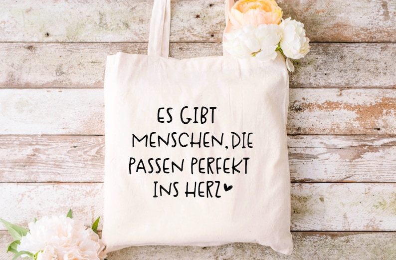 Puede incluir: Una bolsa de tela blanca con texto negro que dice "Es gibt Menschen, die passen perfekt ins Herz" con un peque&ntilde;o s&iacute;mbolo de coraz&oacute;n.