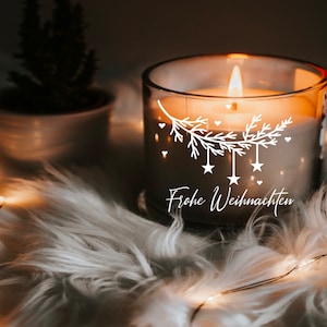 May include: A clear glass candle with a white design of a pine branch with stars and hearts. The candle is lit and the text "Frohe Weihnachten" is written on the side.