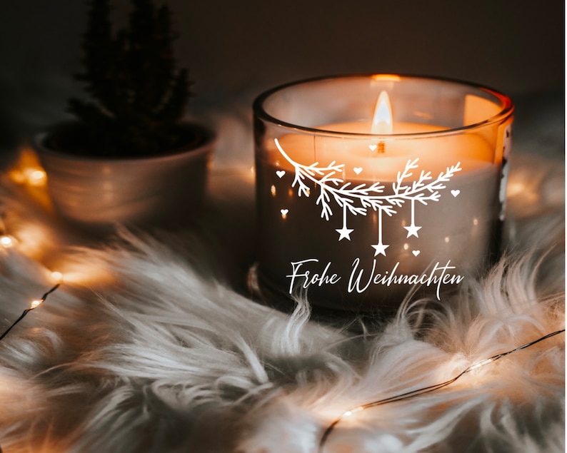 May include: A clear glass candle with a white design of a pine branch with stars and hearts. The candle is lit and the text "Frohe Weihnachten" is written on the side.