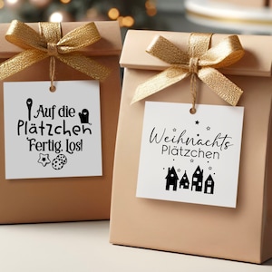 May include: Two brown paper gift bags with gold ribbon and white gift tags. The tag on the left bag says "Auf die Pl&auml;tzchen Fertig. Los!" with a cookie cutter and two cookies. The tag on the right bag says "Weihnachts Pl&auml;tzchen" with a row of gingerbread houses.