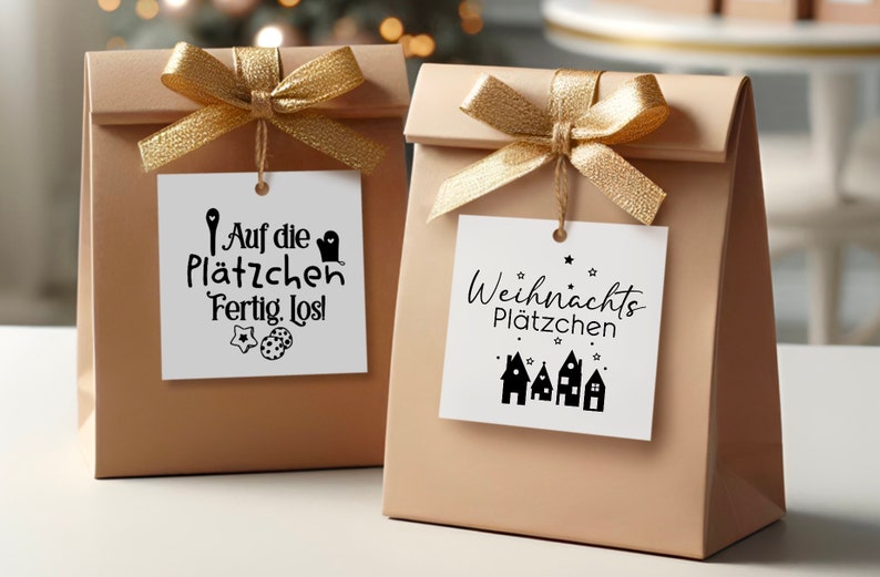 May include: Two brown paper gift bags with gold ribbon and white gift tags. The tag on the left bag says "Auf die Pl&auml;tzchen Fertig. Los!" with a cookie cutter and two cookies. The tag on the right bag says "Weihnachts Pl&auml;tzchen" with a row of gingerbread houses.