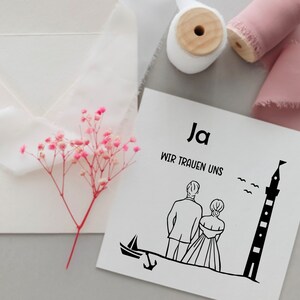 May include: A white card with black text and a black line drawing of a couple standing in front of a lighthouse. The text on the card reads "Ja Wir Trauen Uns".