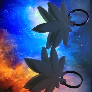 May include: Two white 3D printed marijuana leaf keyrings with silver key rings. The keychains are against a blue and orange galaxy background.