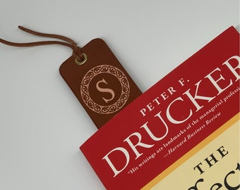 May include: A brown leather bookmark with a gold metal ring and a decorative monogram of the letter "S" in a circle. The bookmark is on top of a red book with the title "Peter F. Drucker" and the text "His writings are landmarks of the managerial profession - Harvard Business Review" printed on the cover.