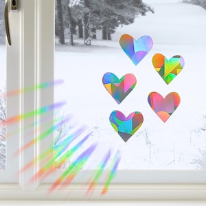Puede incluir: Cinco pegatinas para ventana en forma de corazón con un patrón geométrico holográfico multicolor. Las pegatinas están aplicadas sobre un cristal, proyectando un vibrante destello de luz prismática en la parte inferior izquierda del marco.