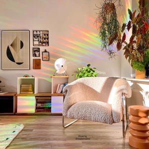Pu&ograve; includere: Una poltrona bianca e soffice con una struttura cromata si trova in una stanza con un effetto di luce prismatica arcobaleno. La stanza ha un pavimento in legno, una libreria con libri e una pianta in vaso.