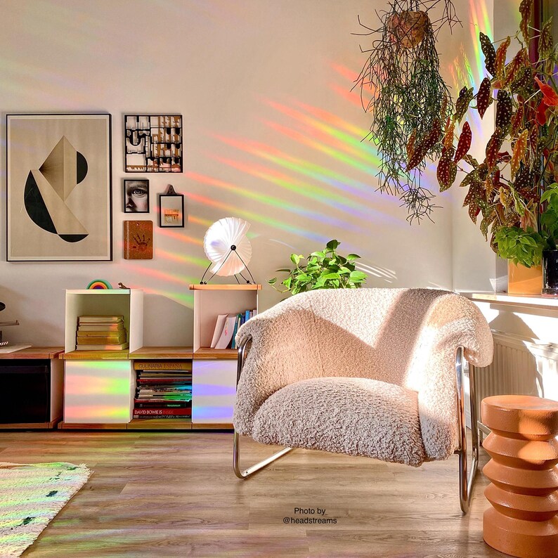 Pu&ograve; includere: Una poltrona bianca e soffice con una struttura cromata si trova in una stanza con un effetto di luce prismatica arcobaleno. La stanza ha un pavimento in legno, una libreria con libri e una pianta in vaso.