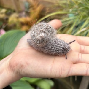 May include: A hand-knitted gray snail with a textured shell. The snail is sitting on a person's hand.