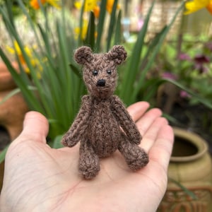 May include: A small brown knitted teddy bear sitting on an open hand. The bear has black eyes and a black nose.