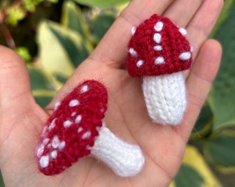 May include: Two knitted mushroom ornaments, one red with white spots and one white. The mushrooms are held in a hand.