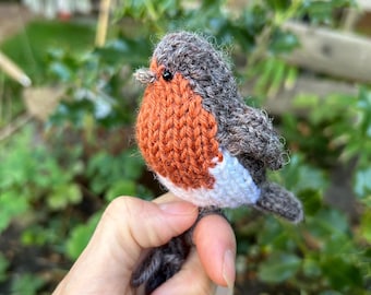 May include: A hand holding a knitted robin bird toy. The bird is brown, orange, and white.