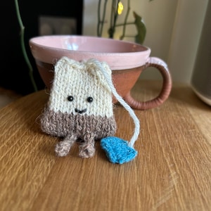Op de afbeelding: Een grillig, handgebreid theezakje met een lachend gezicht, bruine en crèmekleurige kleuren en een blauw label. Het theezakje is geplaatst voor een roze en bruine keramische mok, wat een gezellig, handgemaakt thema creëert.