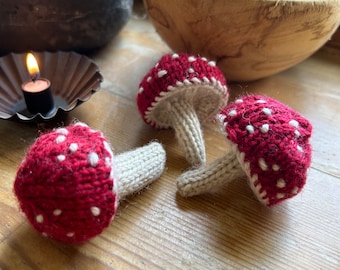 May include: Three knitted red and white mushroom ornaments with white polka dots. The mushrooms have beige stems and are lying on a wooden surface.