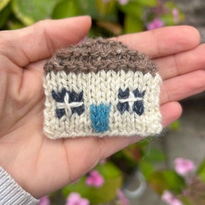 Op de afbeelding: Een miniatuur gebreid huis met een bruin dak, crèmekleurige muren en donkerblauwe raamdetails. De voordeur is helderblauw. Het huis wordt in een hand gehouden en toont het handgemaakte vakmanschap van dit gezellige, decoratieve item.