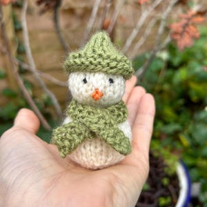 May include: A small knitted snowman with a green hat and scarf. The snowman is sitting in the palm of a hand.