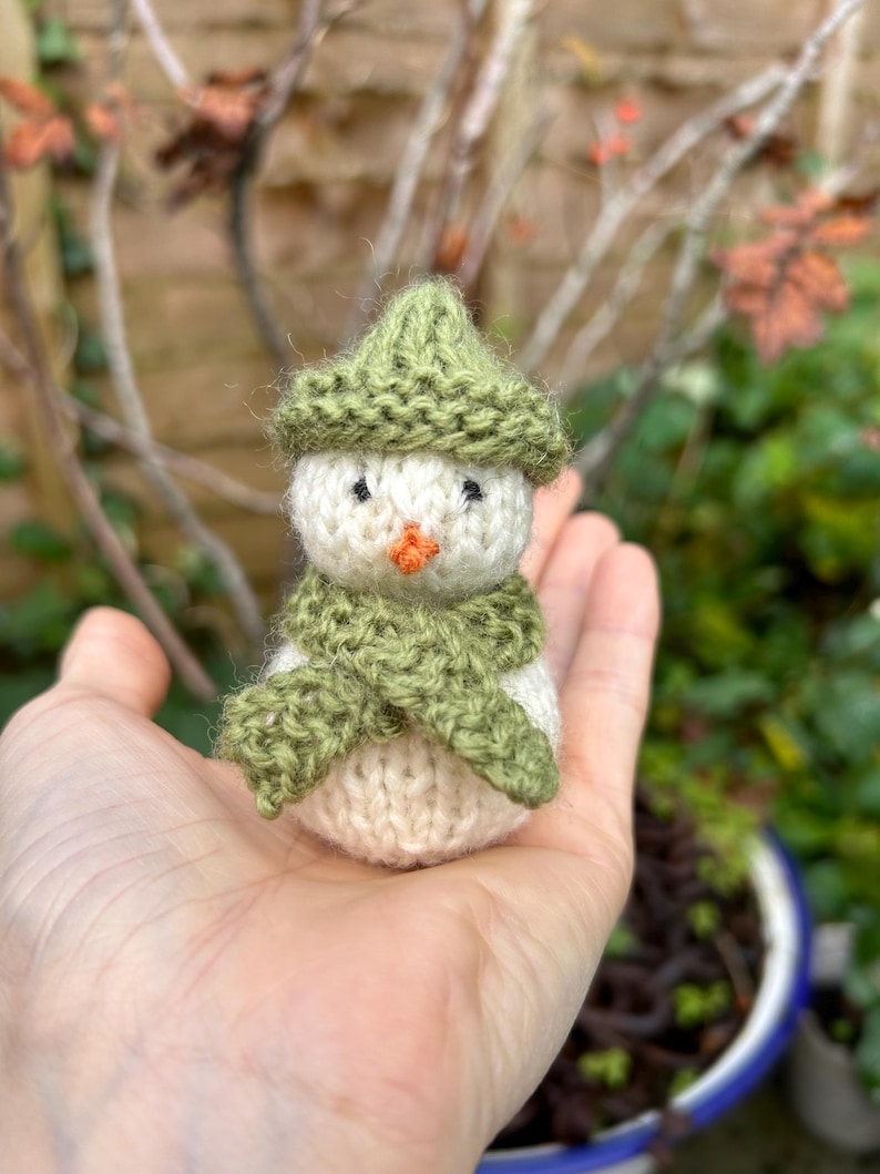 May include: A small knitted snowman with a green hat and scarf. The snowman is sitting in the palm of a hand.