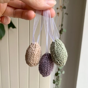 May include: Three knitted Easter eggs in beige, green, and purple, hanging from white ribbon.