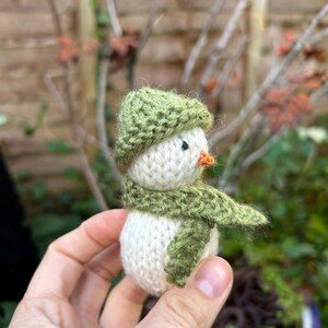 May include: A small knitted snowman with a green hat and scarf. The snowman is white with a small orange nose.