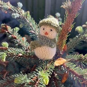 May include: A small knitted snowman ornament with a green hat and scarf, sitting on a branch of a pine tree.