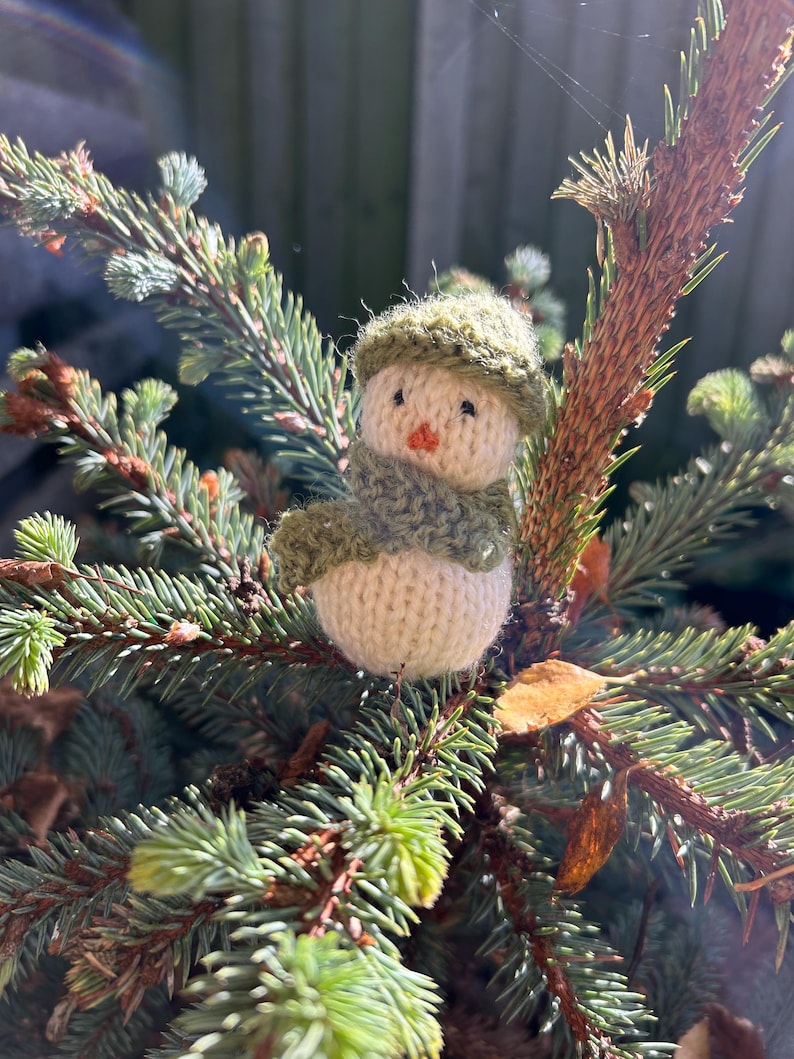 May include: A small knitted snowman ornament with a green hat and scarf, sitting on a branch of a pine tree.