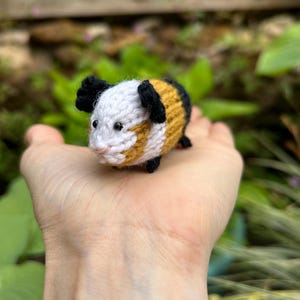 Könnte beinhalten: Eine kleine, handgestrickte Meerschweinchen-Figur. Das Spielzeug hat einen weißen Kopf mit schwarzen Ohren, einen gelb-braunen Körper und schwarze Füße. Das Meerschweinchen wird in einer offenen Hand vor einem Hintergrund aus grünem Laub gehalten.