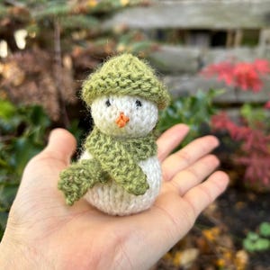 May include: A small knitted snowman wearing a green hat and scarf. The snowman is sitting in the palm of a hand.