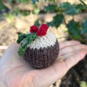 以下が含まれることがあります： 赤いヒイラギの実と緑の葉が付いた、茶色と白のニットのクリスマスプディングのオーナメント