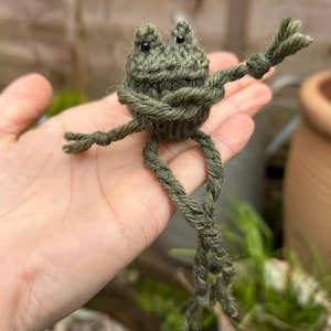 Op de afbeelding: Een groene gebreide kikker met zwarte ogen. De kikker zit op een hand met zijn poten uitgestrekt.