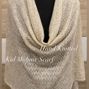 Könnte beinhalten: Handgestrickter, hellbeige Kid-Mohair-Schal. Der Schal ist über einer schwarzen Form drapiert, wodurch das offene Gewebe und die weiche Textur hervorgehoben werden. Der Text "Hand Knitted Kid Mohair Scarf" ist sichtbar.