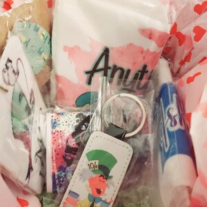 May include: A gift basket filled with various items, including a white bag with the word "Aunt" printed in pink, a blue lip balm, a white keychain with a cartoon image of the Mad Hatter from Alice in Wonderland, and a white and blue patterned fabric.