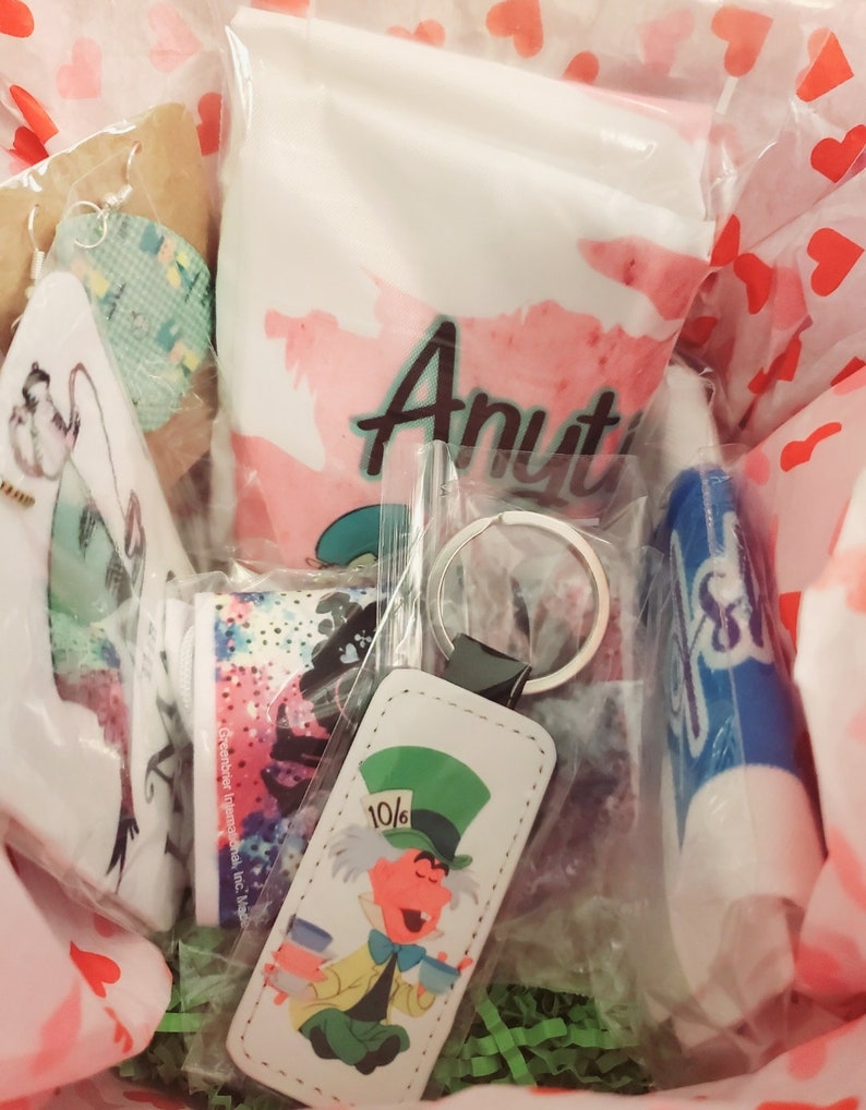May include: A gift basket filled with various items, including a white bag with the word "Aunt" printed in pink, a blue lip balm, a white keychain with a cartoon image of the Mad Hatter from Alice in Wonderland, and a white and blue patterned fabric.