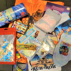May include: A variety of Scooby-Doo themed merchandise including a puzzle, socks, a tumbler, a keychain, and a tote bag. The items are laid out on a brown wooden surface. The tote bag is orange and features the Scooby-Doo logo. The socks are white with a blue and pink tie-dye pattern and feature the Scooby-Doo logo. The tumbler is blue and features a cartoon image of the Mystery Machine. The keychain is a small metal charm with the Scooby-Doo logo. The puzzle is a wooden jigsaw puzzle with a cartoon image of Scooby-Doo and the gang. The tote bag also features the phrase "Ruh Roh" in black letters.