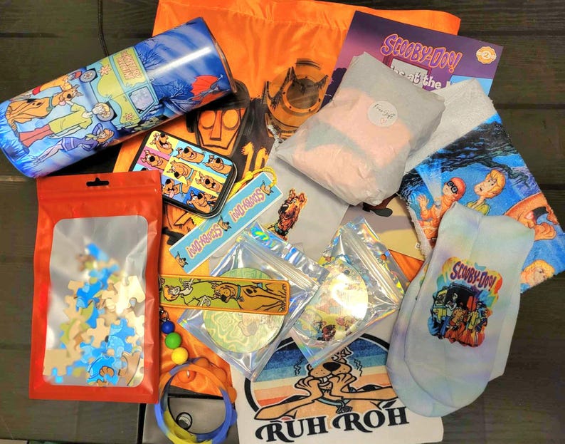 May include: A variety of Scooby-Doo themed merchandise including a puzzle, socks, a tumbler, a keychain, and a tote bag. The items are laid out on a brown wooden surface. The tote bag is orange and features the Scooby-Doo logo. The socks are white with a blue and pink tie-dye pattern and feature the Scooby-Doo logo. The tumbler is blue and features a cartoon image of the Mystery Machine. The keychain is a small metal charm with the Scooby-Doo logo. The puzzle is a wooden jigsaw puzzle with a cartoon image of Scooby-Doo and the gang. The tote bag also features the phrase "Ruh Roh" in black letters.