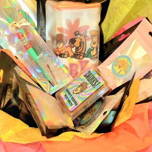 May include: A gift basket filled with various items, including a green and silver pen, a wax melt with the text "Groovy Scented Wax Melts", a sticker with the text "Zainks!" and a Scooby Doo themed bag with the text "Scooby Dooby Doo!"