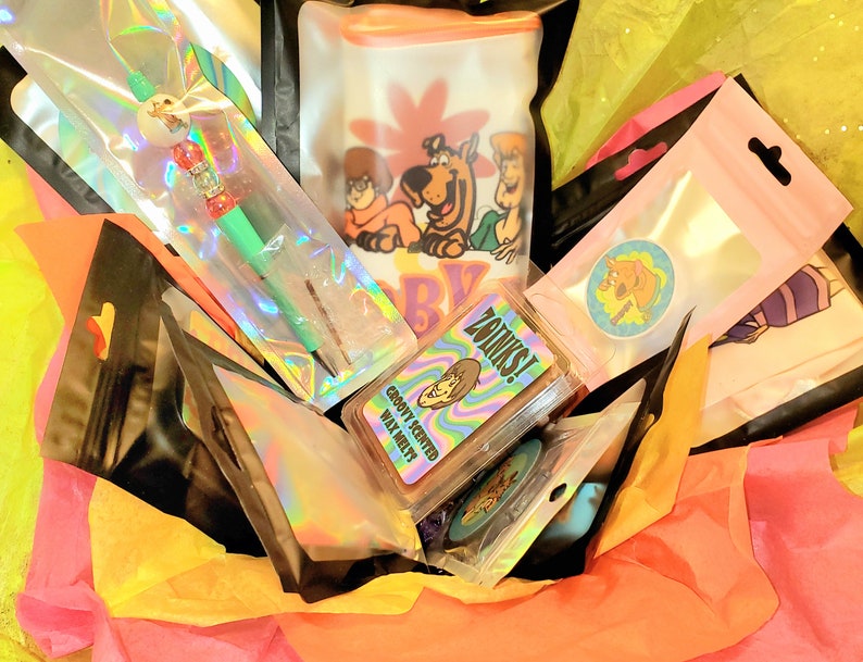 May include: A gift basket filled with various items, including a green and silver pen, a wax melt with the text "Groovy Scented Wax Melts", a sticker with the text "Zainks!" and a Scooby Doo themed bag with the text "Scooby Dooby Doo!"