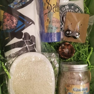 May include: A gift basket filled with bath and body products, including a blue hand sanitizer bottle labeled "Sally Moisturizing Hand Sanitizer", a jar of bath soak labeled "Glowing Frogs Bread", a white loofah, and a pair of black and white earrings.
