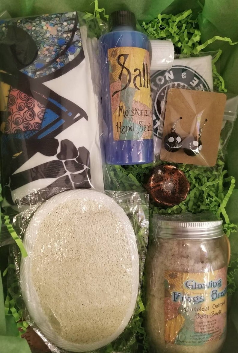 May include: A gift basket filled with bath and body products, including a blue hand sanitizer bottle labeled "Sally Moisturizing Hand Sanitizer", a jar of bath soak labeled "Glowing Frogs Bread", a white loofah, and a pair of black and white earrings.
