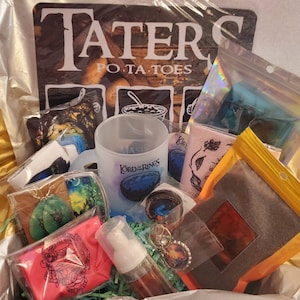 May include: A gift box filled with various items, including a mug with the words "Lord of the Rings" on it, a pink pouch with a graphic design, a small bag of brown powder, a pair of green earrings, and a keychain with a star-shaped charm. The box also includes a sign that says "Tater Po-ta-toes".