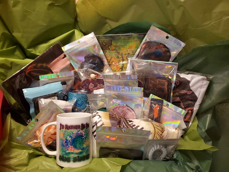 May include: A gift box filled with various items, including a white mug with the text "I'd Rather Be In The Shire", a map of Middle-earth, and a variety of other fantasy-themed items.
