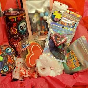 May include: A pink gift box filled with various items, including a black and white tumbler with a spooky character design, a heart-shaped cookie, a pink heart-shaped hair clip, a small bag of bath salts, and a clear bag with a colorful beaded necklace.