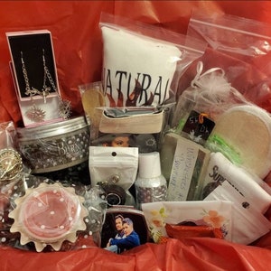 May include: A gift basket filled with various items, including earrings, a jar labeled "BURN", a bag of bath salts with "NATURAL" printed on it, soap, and a photo. The items are arranged on red tissue paper.