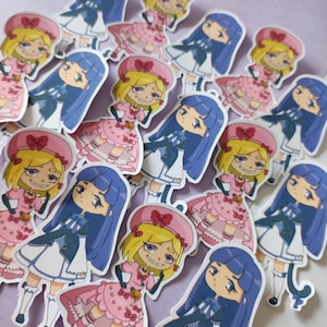 Könnte beinhalten: Eine Sammlung von Aufklebern mit zwei verschiedenen Anime-Charakteren. Ein Charakter hat blaue Haare und ein weißes Kleid mit einer schwarzen Schleife, während der andere blonde Haare und ein rosa Kleid mit einer roten Schleife hat. Beide Charaktere haben einen niedlichen und fantasievollen Stil.