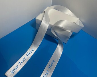 May include: Two white satin ribbons with the text "Your Text Here" printed in blue on each ribbon. The ribbons are tied around a white box.
