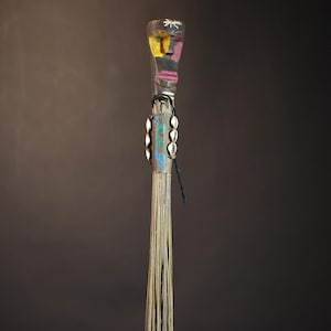 African traditional fly whisk with long plant fiber tassels, a carved wooden handle featuring a painted tribal face, and cowrie shell accents. Handcrafted ceremonial whip tool used in African rituals, displayed against a dark background. Vintage wall art or decor piece reflecting authentic African heritage and craftsmanship.