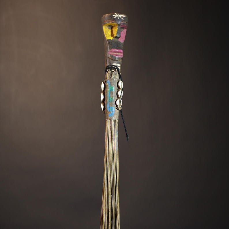 African traditional fly whisk with long plant fiber tassels, a carved wooden handle featuring a painted tribal face, and cowrie shell accents. Handcrafted ceremonial whip tool used in African rituals, displayed against a dark background. Vintage wall art or decor piece reflecting authentic African heritage and craftsmanship.