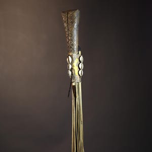 African traditional fly whisk with long plant fiber tassels, a carved wooden handle featuring a painted tribal face, and cowrie shell accents. Handcrafted ceremonial whip tool used in African rituals, displayed against a dark background. Vintage wall art or decor piece reflecting authentic African heritage and craftsmanship.