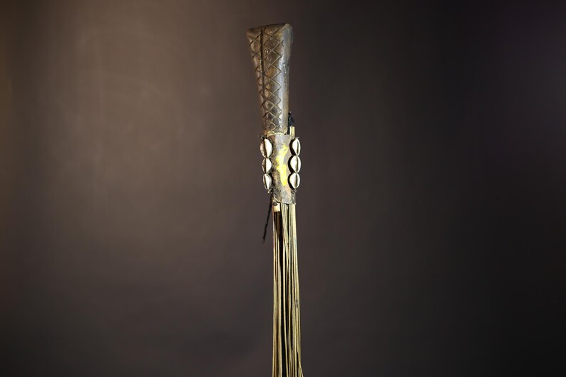 African traditional fly whisk with long plant fiber tassels, a carved wooden handle featuring a painted tribal face, and cowrie shell accents. Handcrafted ceremonial whip tool used in African rituals, displayed against a dark background. Vintage wall art or decor piece reflecting authentic African heritage and craftsmanship.