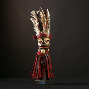 Könnte beinhalten: Eine Holzfigur mit einem rot-schwarzen Federkopfschmuck und einer rot-schwarzen gemusterten Robe. Die Figur hat ein schwarzes Gesicht mit weißen Augen und einem weißen Mund.